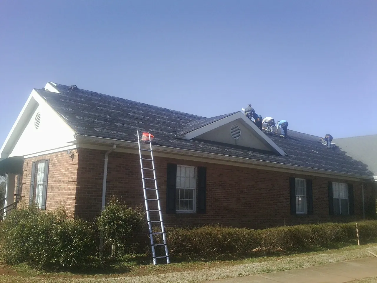 Crew tearing shingles off a brick ranch roof with a ladder set — Metal Roof Repair work by our crew in Devens