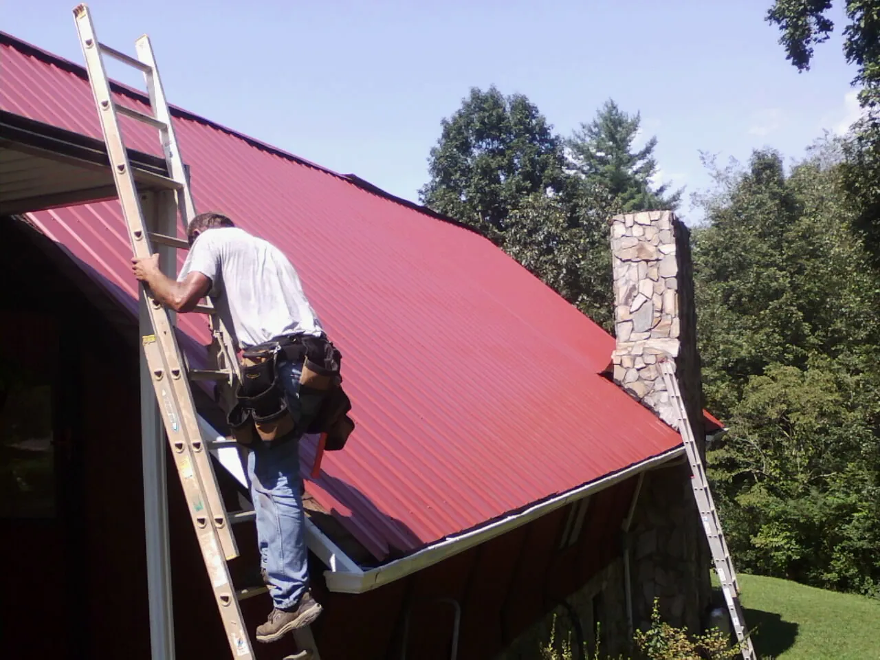 Roofer on a ladder beside a red metal roof and stone chimney — Soffit & Fascia Repair by Roofing Brothers in Devens